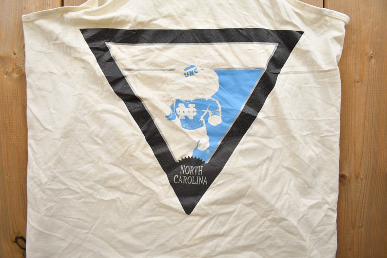 May include: White tank top with a black triangle graphic featuring a cartoon mascot wearing a blue and white jersey with the text "UNC" on the hat and "NORTH CAROLINA" below the graphic.