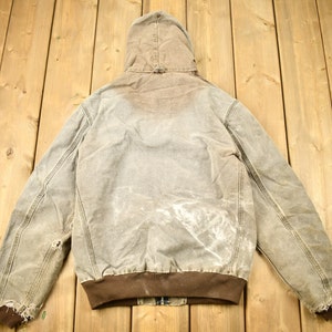 Może przedstawiać: Brązowa kurtka robocza z kapturem o wyglądzie zużytym. Kurtka ma brązowy kołnierz z dzianiny typu corduroy i brązowe ściągacze. Kurtka jest wykonana z grubego, wytrzymałego materiału.