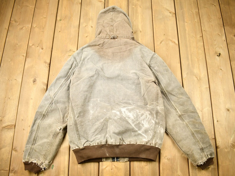 Może przedstawiać: Brązowa kurtka robocza z kapturem o wyglądzie zużytym. Kurtka ma brązowy kołnierz z dzianiny typu corduroy i brązowe ściągacze. Kurtka jest wykonana z grubego, wytrzymałego materiału.