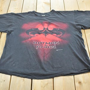 May include: A dark grey t-shirt with a red and black graphic of the Batman logo and the words "BATMAN & ROBIN" in red. The shirt is made of a soft material and has short sleeves.