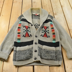 Op de afbeelding: Een grijs gebreid vest met een sjaalkraag, lange mouwen met boorden en een knoopsluiting. Het kledingstuk heeft een geometrisch patroon in rood, zwart en groenblauw, twee voorzakken en een onderste gedeelte van gestructureerd grijs breiwerk.