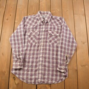 Puede incluir: Una camisa de manga larga a cuadros con cuello abotonado y dos bolsillos en el pecho. La camisa es blanca con un patrón de cuadros rojos y azules.