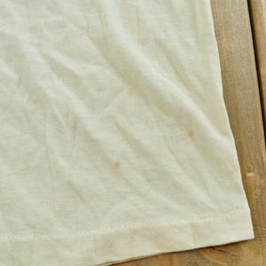 May include: A plain off-white t-shirt featuring a soft, lightweight fabric with visible wrinkles and two small, light brown circular stains located on the lower right side of the garment, near the hemline, showing a simple, casual construction.