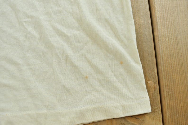 May include: A plain off-white t-shirt featuring a soft, lightweight fabric with visible wrinkles and two small, light brown circular stains located on the lower right side of the garment, near the hemline, showing a simple, casual construction.