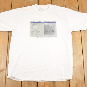 以下が含まれることがあります： グラフィックプリントが施された白いTシャツ。「Landsat Archive Conversion System」と「LACS Integration System」の文字と、図と風景画像が含まれています。柔らかい素材で作られたTシャツです。