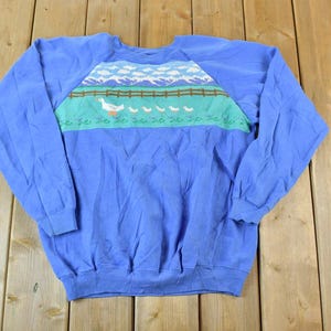 Puede incluir: Una sudadera azul de manga larga con un gr&aacute;fico colorido de un ganso y gansitos caminando sobre una valla frente a una cordillera.