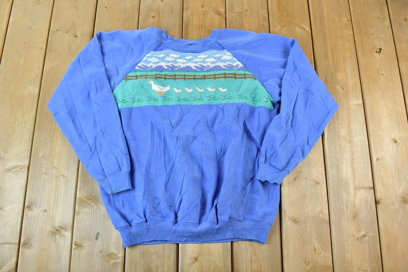 Puede incluir: Una sudadera azul de manga larga con un gr&aacute;fico colorido de un ganso y gansitos caminando sobre una valla frente a una cordillera.