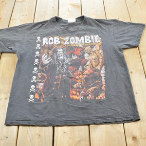 Puede incluir: Una camiseta gris oscuro con un gráfico de Rob Zombie y otras figuras de terror. La camiseta tiene el texto "Rob Zombie" y "Hellbilly Deluxe" impreso en la parte delantera, con un diseño de calavera y huesos cruzados en el lado izquierdo.