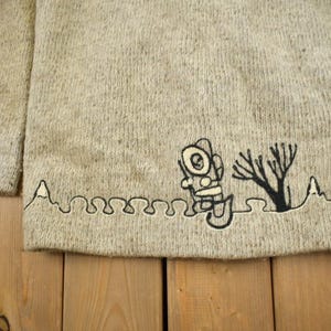 May include: A light beige sweater with a crew neck and long sleeves. The bottom hem features a black embroidered design of a diver, a tree, and a wavy line. The sweater is made of a textured knit fabric.