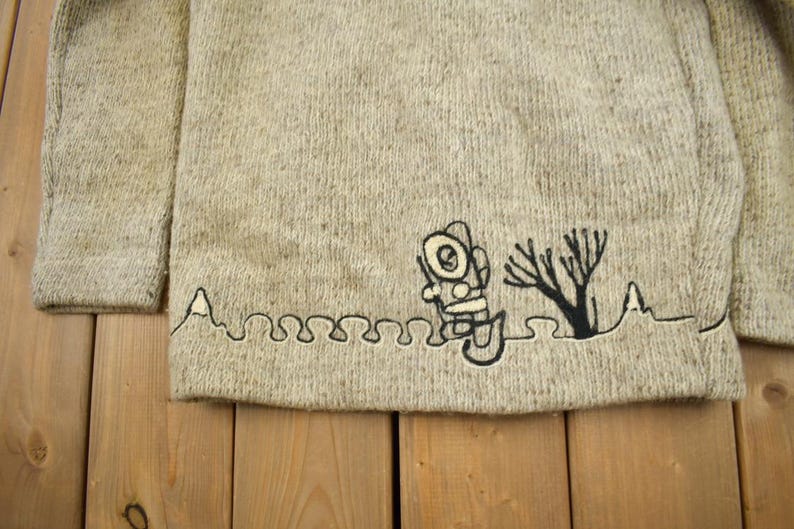 May include: A light beige sweater with a crew neck and long sleeves. The bottom hem features a black embroidered design of a diver, a tree, and a wavy line. The sweater is made of a textured knit fabric.