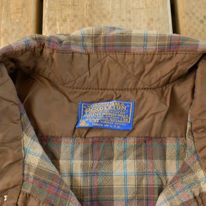 Puede incluir: Primer plano del cuello de una camisa de cuadros vintage. La camisa presenta un forro acolchado marr&oacute;n y una etiqueta azul con texto blanco. El estampado de cuadros incluye tonos de marr&oacute;n, azul y rojo. Hecho en EE. UU.