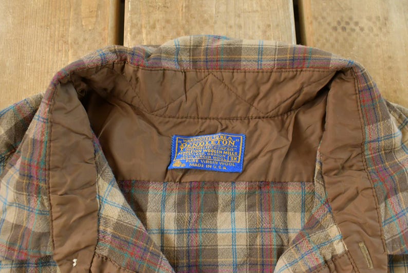 Puede incluir: Primer plano del cuello de una camisa de cuadros vintage. La camisa presenta un forro acolchado marr&oacute;n y una etiqueta azul con texto blanco. El estampado de cuadros incluye tonos de marr&oacute;n, azul y rojo. Hecho en EE. UU.