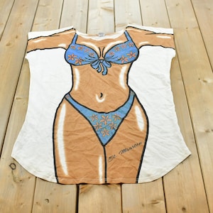 Pode incluir: Uma t-shirt branca com uma impressão gráfica de um torso bronzeado a usar um biquíni azul com padrões florais cor de laranja. O design inclui o texto St. Maarten impresso em letra preta no lado inferior direito da cueca do biquíni.