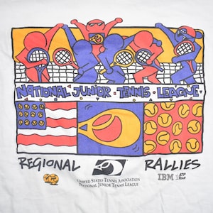 Puede incluir: Una camiseta blanca con una ilustraci&oacute;n de dibujos animados de ni&ntilde;os jugando al tenis. La ilustraci&oacute;n incluye una red de tenis, raquetas y pelotas de tenis. El texto "NATIONAL JUNIOR TENNIS LEAGUE" est&aacute; impreso sobre la ilustraci&oacute;n. El texto "REGIONAL" est&aacute; impreso debajo de la ilustraci&oacute;n a la izquierda. El texto "RALLIES" est&aacute; impreso debajo de la ilustraci&oacute;n a la derecha. El texto "IBM ATP Tour" est&aacute; impreso debajo del texto "RALLIES". El texto "UNITED STATES TENNIS ASSOCIATION NATIONAL JUNIOR TENNIS LEAGUE" est&aacute; impreso debajo del texto "RALLIES" y encima del texto "IBM ATP Tour".