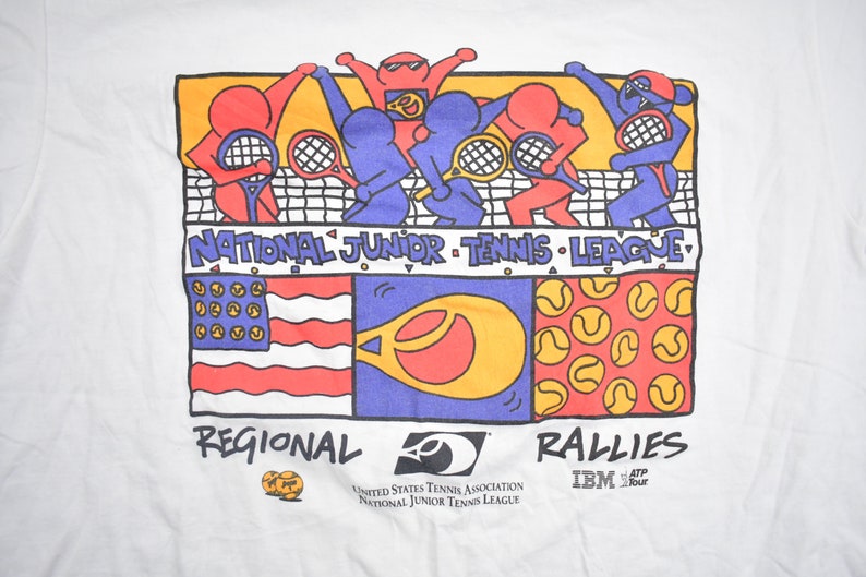Puede incluir: Una camiseta blanca con una ilustraci&oacute;n de dibujos animados de ni&ntilde;os jugando al tenis. La ilustraci&oacute;n incluye una red de tenis, raquetas y pelotas de tenis. El texto "NATIONAL JUNIOR TENNIS LEAGUE" est&aacute; impreso sobre la ilustraci&oacute;n. El texto "REGIONAL" est&aacute; impreso debajo de la ilustraci&oacute;n a la izquierda. El texto "RALLIES" est&aacute; impreso debajo de la ilustraci&oacute;n a la derecha. El texto "IBM ATP Tour" est&aacute; impreso debajo del texto "RALLIES". El texto "UNITED STATES TENNIS ASSOCIATION NATIONAL JUNIOR TENNIS LEAGUE" est&aacute; impreso debajo del texto "RALLIES" y encima del texto "IBM ATP Tour".