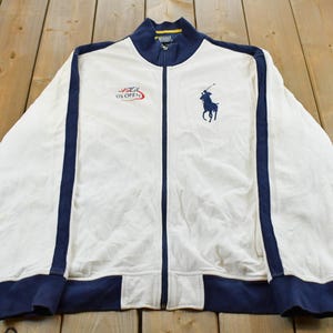 Pode incluir: Um casaco de treino branco com fecho de correr e detalhes em azul-marinho na gola, punhos, bainha e mangas. O peito apresenta um logótipo de pónei Polo Ralph Lauren bordado em azul-marinho à esquerda e o logótipo do US Open à direita.