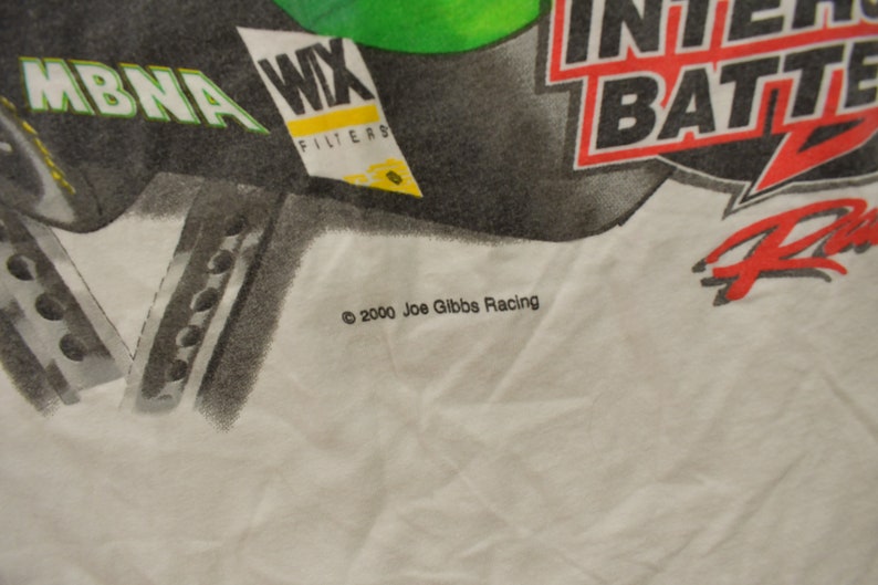 May include: White t-shirt featuring a black and green graphic of a car with the text "MBNA", "WIX Filters", "Interstate Batteries", and "Racing". The text "&copy; 2000 Joe Gibbs Racing" is printed at the bottom of the shirt.