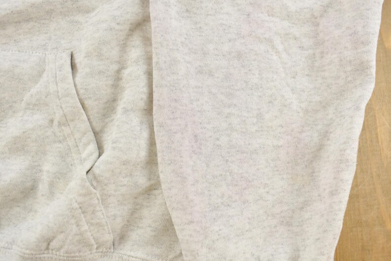 Op de afbeelding: Een lichtgrijs gem&ecirc;leerd sweatshirt met een zachte, gespikkelde textuur. Het kledingstuk is voorzien van een zichtbare voorzak met gestikte naden, wat het gebreide materiaal en het subtiele gevlekte patroon van de stof benadrukt.