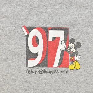 May include: A gray t-shirt featuring a graphic of Mickey Mouse next to the number "97" in red and white, with "Walt Disney World" printed below. Mickey is wearing red shorts and yellow shoes.