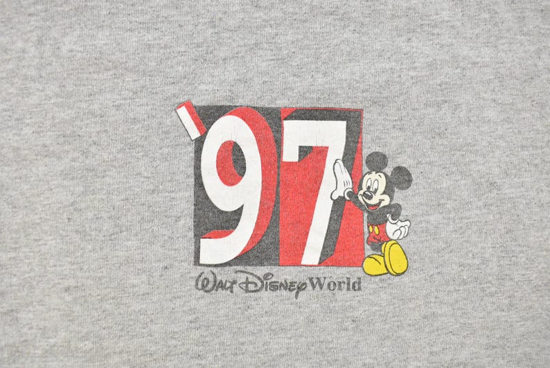 May include: A gray t-shirt featuring a graphic of Mickey Mouse next to the number "97" in red and white, with "Walt Disney World" printed below. Mickey is wearing red shorts and yellow shoes.