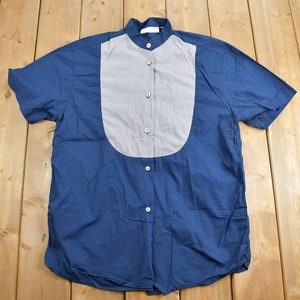 Könnte beinhalten: Ein blaues Kurzarmhemd mit einem weißen und grauen gestreiften Einsatz vorne. Das Hemd hat einen Stehkragen und einen Knopfverschluss.