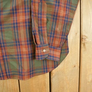 May include: Close-up of a button-down shirt with a plaid pattern in shades of green, orange, and blue. The shirt has a buttoned cuff and a classic collar. The fabric appears to be a woven material.