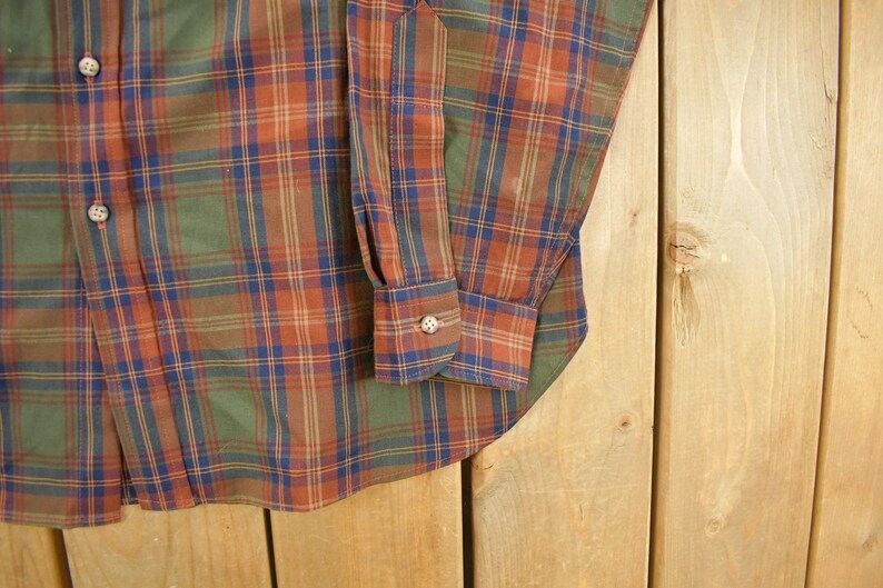 May include: Close-up of a button-down shirt with a plaid pattern in shades of green, orange, and blue. The shirt has a buttoned cuff and a classic collar. The fabric appears to be a woven material.