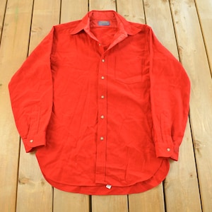 May include: A bright red button-down shirt with long sleeves, a collar, and a chest pocket. The shirt is made of a soft fabric and has gold-colored buttons. The shirt is laid flat on a wooden surface.
