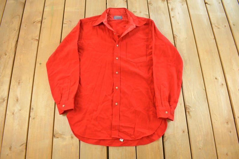 May include: A bright red button-down shirt with long sleeves, a collar, and a chest pocket. The shirt is made of a soft fabric and has gold-colored buttons. The shirt is laid flat on a wooden surface.