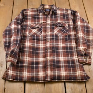 Pode incluir: Uma camisa de flanela xadrez marrom, preto e branco com mangas compridas e fechamento com botões.