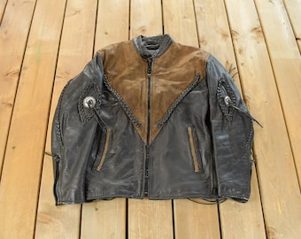 May include: Black leather jacket with a brown suede yoke and a zip-up front. The jacket features braided accents, two front pockets, and decorative metal details on the shoulders. A vintage biker-style jacket.