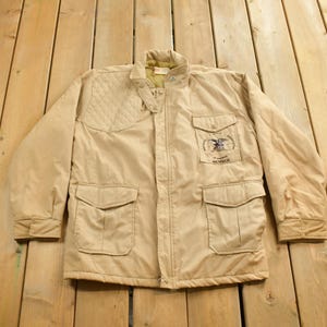 Puede incluir: Una chaqueta acolchada beige con cierre de cremallera y dos bolsillos grandes. La chaqueta tiene un parche en el pecho izquierdo con el texto "America Member".