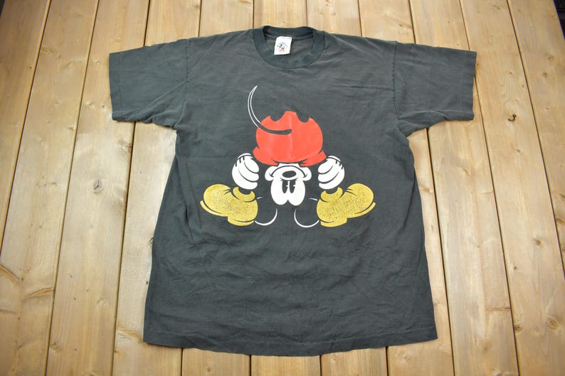 May include: Black t-shirt with a graphic of Mickey Mouse in a red hat and yellow shoes. The graphic shows Mickey Mouse with his hands on his head, looking down.