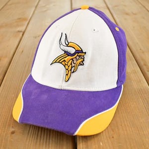 Può includere: Un cappellino da baseball viola, bianco e giallo con il logo dei Minnesota Vikings. Il cappellino ha un pannello frontale bianco con il logo della squadra, una visiera e pannelli laterali viola e dettagli gialli.