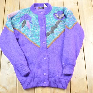 May include: Purple cardigan sweater with a colorful, geometric pattern and a button closure. The sweater has a textured, fuzzy look.