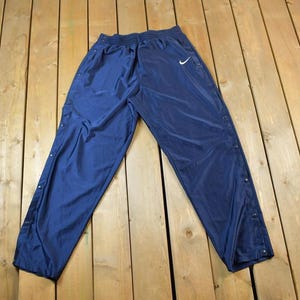 May include: Navy blue track pants with a white Nike swoosh logo and snap buttons down the sides.