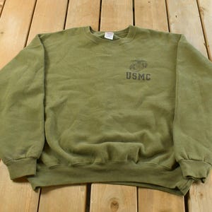 May include: Olive green crewneck sweatshirt with the text "USMC" printed on the upper left chest. The sweatshirt has ribbed cuffs and a ribbed hem. The fabric appears to be a soft, comfortable material.