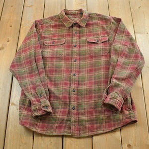 Puede incluir: Una camisa de franela abotonada de manga larga con un estampado de cuadros rojos, verdes y beige. La camisa tiene dos bolsillos en el pecho y puños abotonados. La camisa está tendida sobre una superficie de madera.