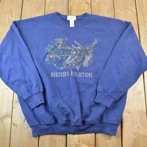 Op de afbeelding: Een blauw sweatshirt met een afbeelding van een eland en de tekst "Northern Reflections" gedrukt op de voorkant.