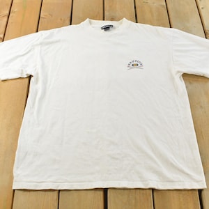 May include: A white Nautica t-shirt with a crew neck. The shirt features a small logo on the upper left chest. The logo includes the word "Nautica" above a graphic of a sailboat and oars.