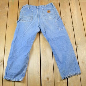 K&ouml;nnte beinhalten: Hellblaue Jeans-Arbeitshose mit mehreren Taschen, einschlie&szlig;lich einer Hammerschlaufe. Die Hose hat einen braunen Lederaufn&auml;her auf der Ges&auml;&szlig;tasche und liegt auf einer Holzoberfl&auml;che.