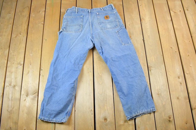 K&ouml;nnte beinhalten: Hellblaue Jeans-Arbeitshose mit mehreren Taschen, einschlie&szlig;lich einer Hammerschlaufe. Die Hose hat einen braunen Lederaufn&auml;her auf der Ges&auml;&szlig;tasche und liegt auf einer Holzoberfl&auml;che.