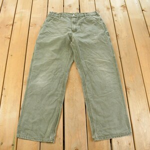 Puede incluir: Pantalones de trabajo verde oliva con cierre de bot&oacute;n y m&uacute;ltiples bolsillos. Los pantalones est&aacute;n hechos de una tela duradera y tienen un dise&ntilde;o de pierna recta. Los pantalones est&aacute;n colocados sobre una superficie de madera.