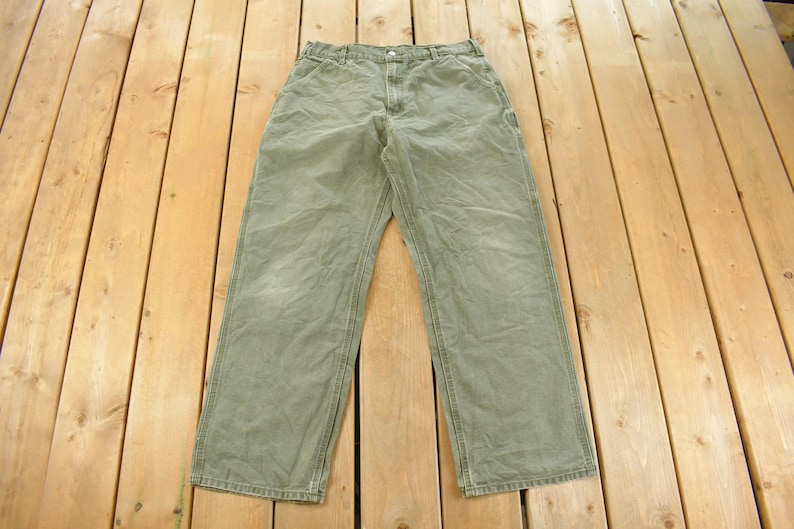 Puede incluir: Pantalones de trabajo verde oliva con cierre de bot&oacute;n y m&uacute;ltiples bolsillos. Los pantalones est&aacute;n hechos de una tela duradera y tienen un dise&ntilde;o de pierna recta. Los pantalones est&aacute;n colocados sobre una superficie de madera.