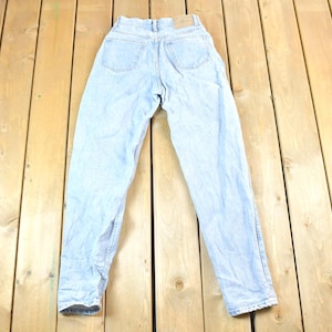 Puede incluir: Vaqueros de mezclilla azul claro con cintura alta, bolsillos traseros visibles y un parche de cuero. Los vaqueros están colocados sobre una superficie de madera, mostrando su lavado descolorido y diseño clásico. Prenda vintage.