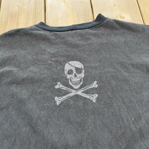 May include: Dark gray t-shirt featuring a white skull and crossbones graphic. The skull has an eye patch. The shirt has a dark blue neckline.