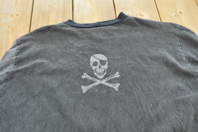 May include: Dark gray t-shirt featuring a white skull and crossbones graphic. The skull has an eye patch. The shirt has a dark blue neckline.