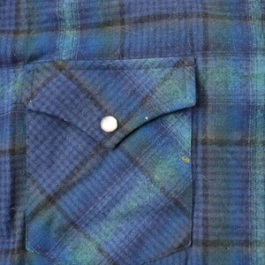 May include: Close-up of a blue and green plaid flannel shirt. The shirt has a pocket on the left side with a silver snap closure. Additional silver snap closures are visible on the right side.