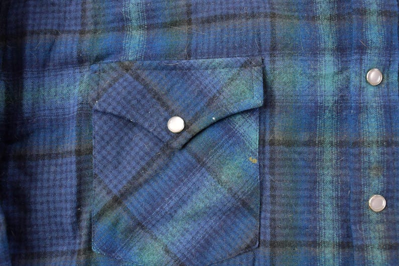May include: Close-up of a blue and green plaid flannel shirt. The shirt has a pocket on the left side with a silver snap closure. Additional silver snap closures are visible on the right side.