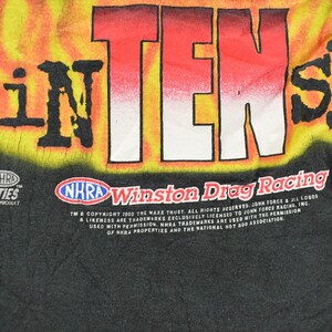 Op de afbeelding: Zwart T-shirt met een opvallende afbeelding van "iNTENSE" in rode en witte letters tegen een gele en oranje vlammenachtergrond. Voorzien van "NHRA Winston Drag Racing" branding.